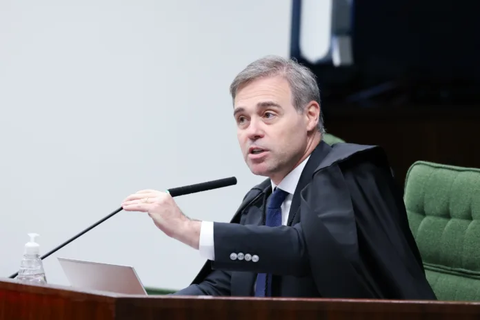 Ministro André Mendonça, do STF, durante sessão no plenário, trajando toga e gravata, gesticulando enquanto se pronuncia. À frente, estão um microfone, um laptop e um frasco de álcool em gel.