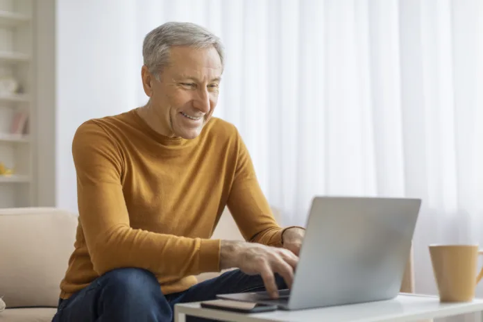 Aposentado sorridente usando um laptop em casa. Imagem ilustrativa para matéria sobre consulta e pagamento de atrasados do INSS liberados pela Justiça