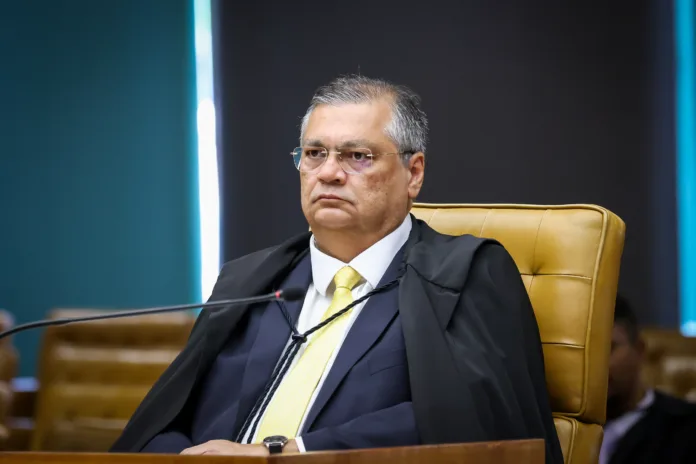 Ministro Flávio Dino, do Supremo Tribunal Federal (STF), sentado em uma cadeira de couro no plenário, trajando toga e gravata amarela, com expressão séria durante a análise de processos.