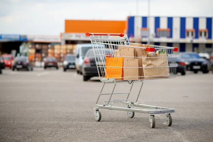 Carrinho de supermercado com compras no estacionamento, com veículos e fachada do mercado ao fundo. Ilustra matéria sobre decisão judicial que responsabiliza supermercado por danos em veículo no estacionamento.