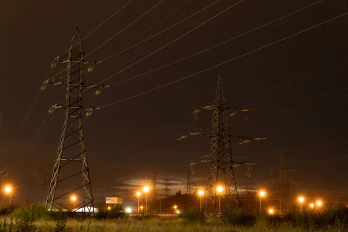 Torres de transmissão de energia elétrica iluminadas à noite, com fios de alta tensão e postes de luz ao fundo. Representação do setor energético e infraestrutura elétrica.