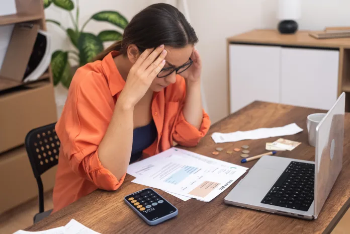 Mulher preocupada analisa contas e boletos bancários em frente ao notebook, representando problemas financeiros e cobrança indevida.