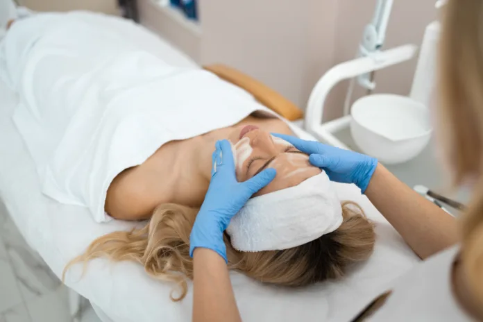 Profissional com luvas realiza procedimento estético facial em paciente deitada em clínica de estética.