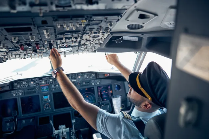 Piloto dentro da cabine de comando de um avião ajustando os controles superiores, representando a atuação profissional na aviação.