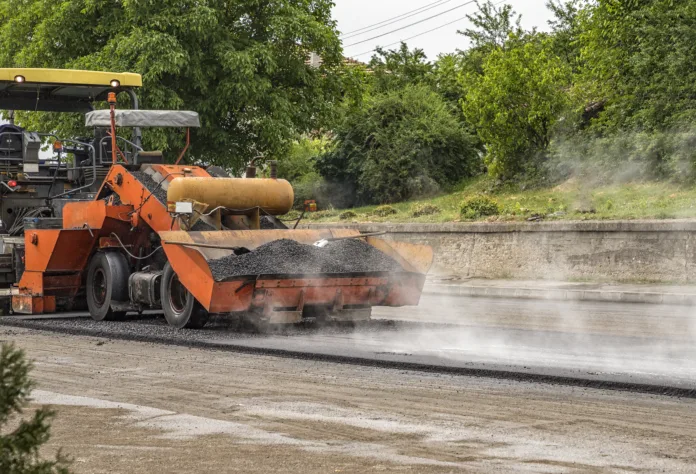 Máquina de pavimentação aplicando asfalto quente em uma rua, com vapor subindo devido à alta temperatura do material. A imagem ilustra obras de pavimentação urbana, contexto de um caso judicial sobre indenização por danos causados a uma residência.