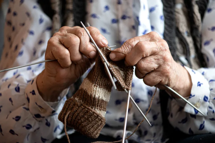 Mãos idosas tricotando um par de meias de lã em tons marrons com agulhas de tricô. Representação da rotina e bem-estar da terceira idade.