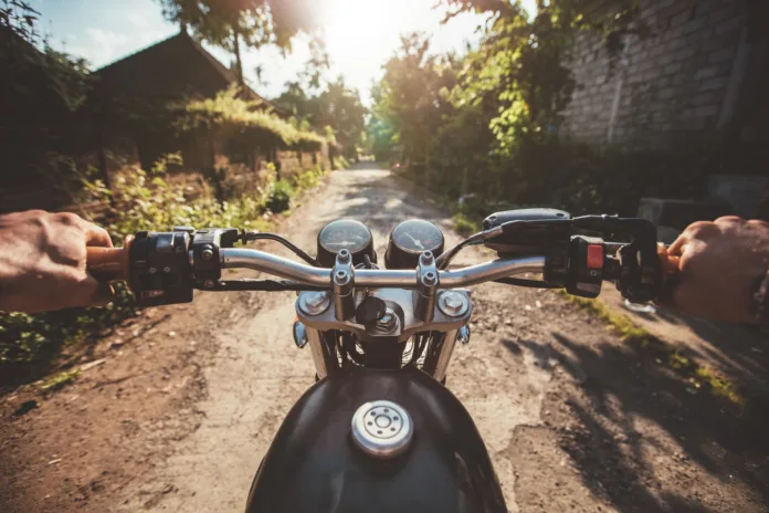 Visão em primeira pessoa de motociclista pilotando moto clássica em estrada de terra, com sol brilhando ao fundo e vegetação ao redor.