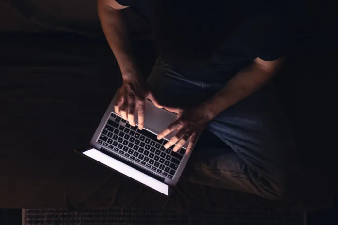 Homem digitando em um notebook no escuro, iluminado apenas pela tela do dispositivo. Representação da dificuldade de trabalho durante uma queda de energia.