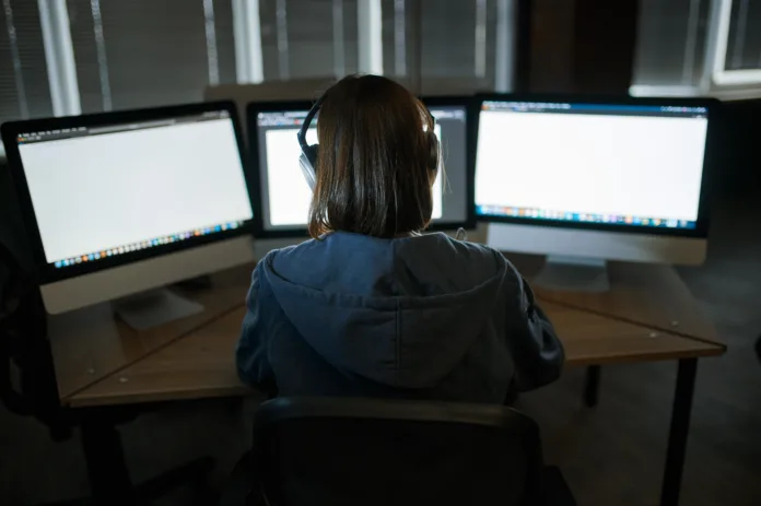 Pessoa de capuz e fones de ouvido sentada em frente a três monitores de computador com telas em branco, em um ambiente escuro. Imagem ilustrativa de atividade digital e monitoramento financeiro.