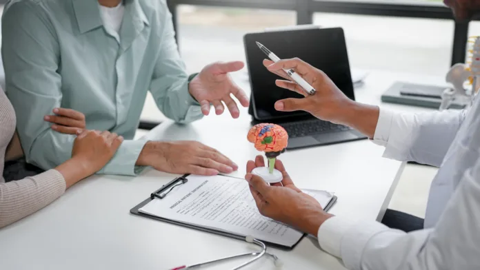 Consulta médica com neurologista: médico explica diagnóstico a pacientes utilizando modelo de cérebro. Documentos e estetoscópio sobre a mesa indicam avaliação clínica.