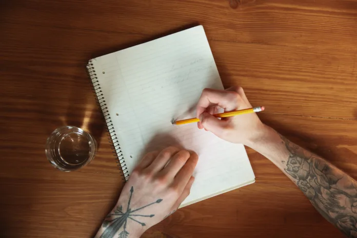 Mãos tatuadas escrevendo com lápis em caderno espiral sobre mesa de madeira, ao lado de um copo de água. Representação da produção literária no cárcere.